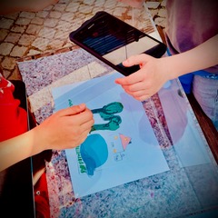 2 Kinder-Hände legen eine Figur aus Papier auf ein Blatt. Darüber hängt ein Tablet und filmt.