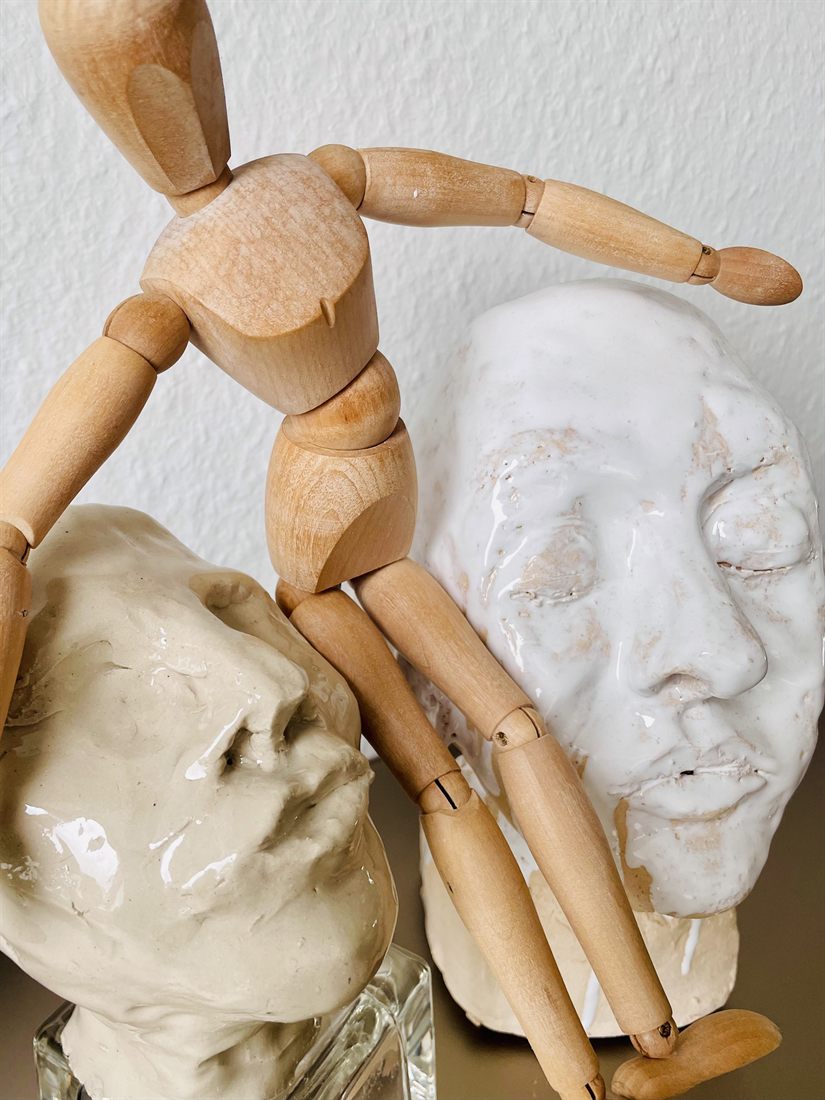 Eine hölzerne Gliederpuppe lehnt zwischen zwei modellierten Köpfen: links ein beiger Tonkopf, rechts ein glänzend weiß glasierter Kopf mit geschlossenen Augen.