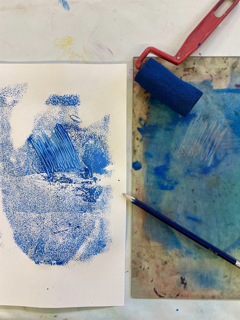 Links ein blauer Abdruck auf weißem Papier, rechts die eingefärbte Druckplatte mit den Spuren einer Zeichnung. Darauf eine rote Farbwalze mit blauer Rolle und ein Bleistift.