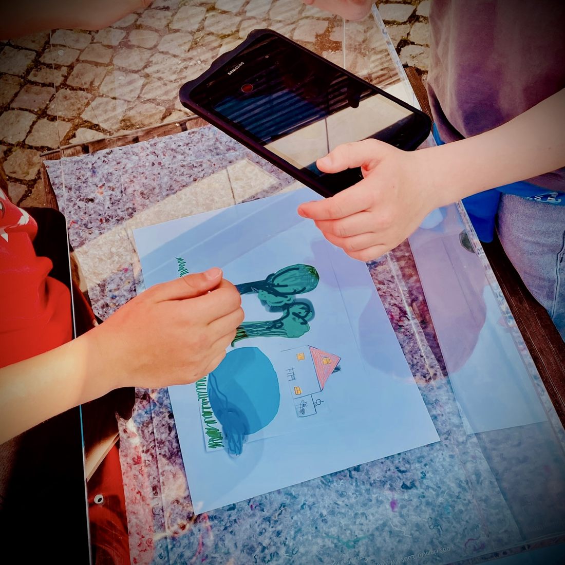 Zwei Kinder an einem Tisch im Freien: eine Hand hält ein Tablet über ein gemaltes Bild mit Haus und Bäumen, eine andere Hand schiebt eine ausgeschnittene Baumfigur über das Blatt.