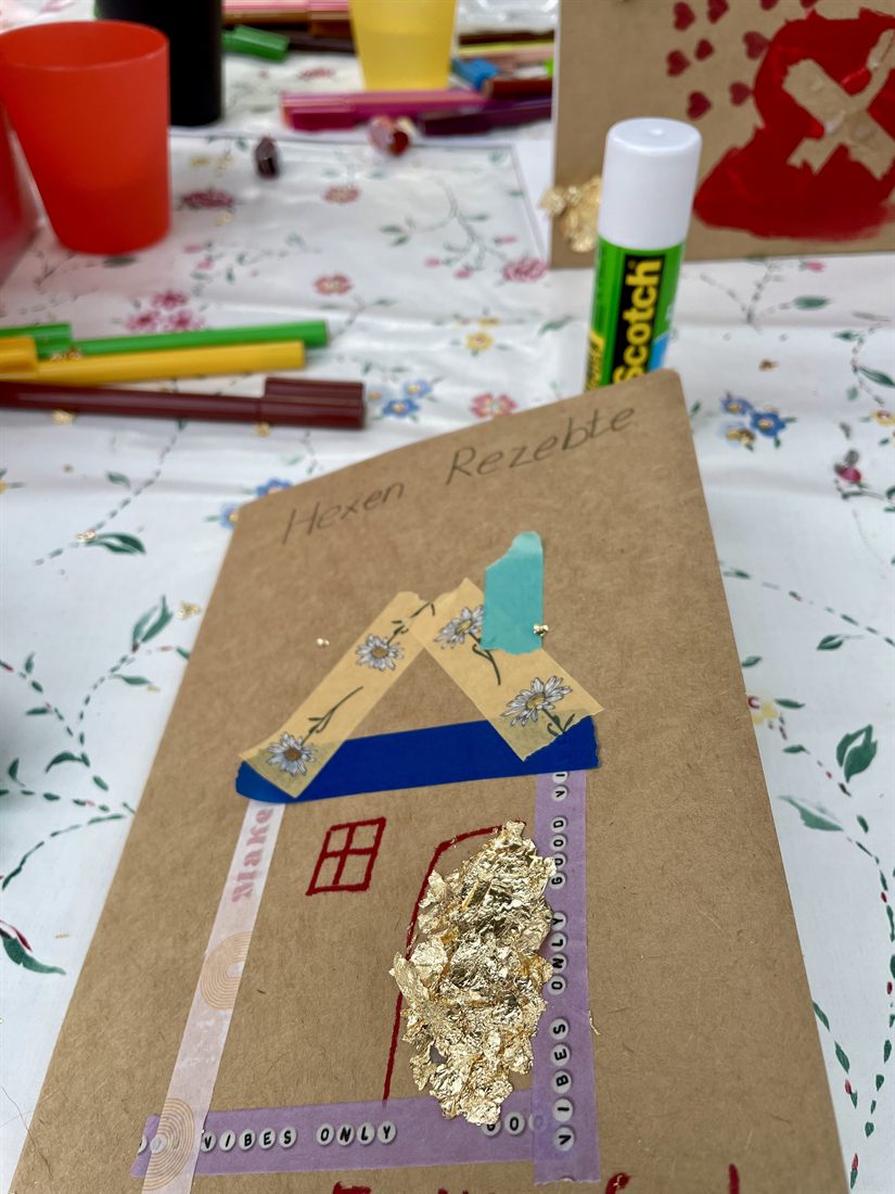 Ein selbst gestaltetes Heft aus braunem Papier mit der Aufschrift „Hexen Rezebte“, darauf ein Haus aus Klebeband, buntem Papier und Blattgold. Daneben Filzstifte, Becher und ein Klebestift.