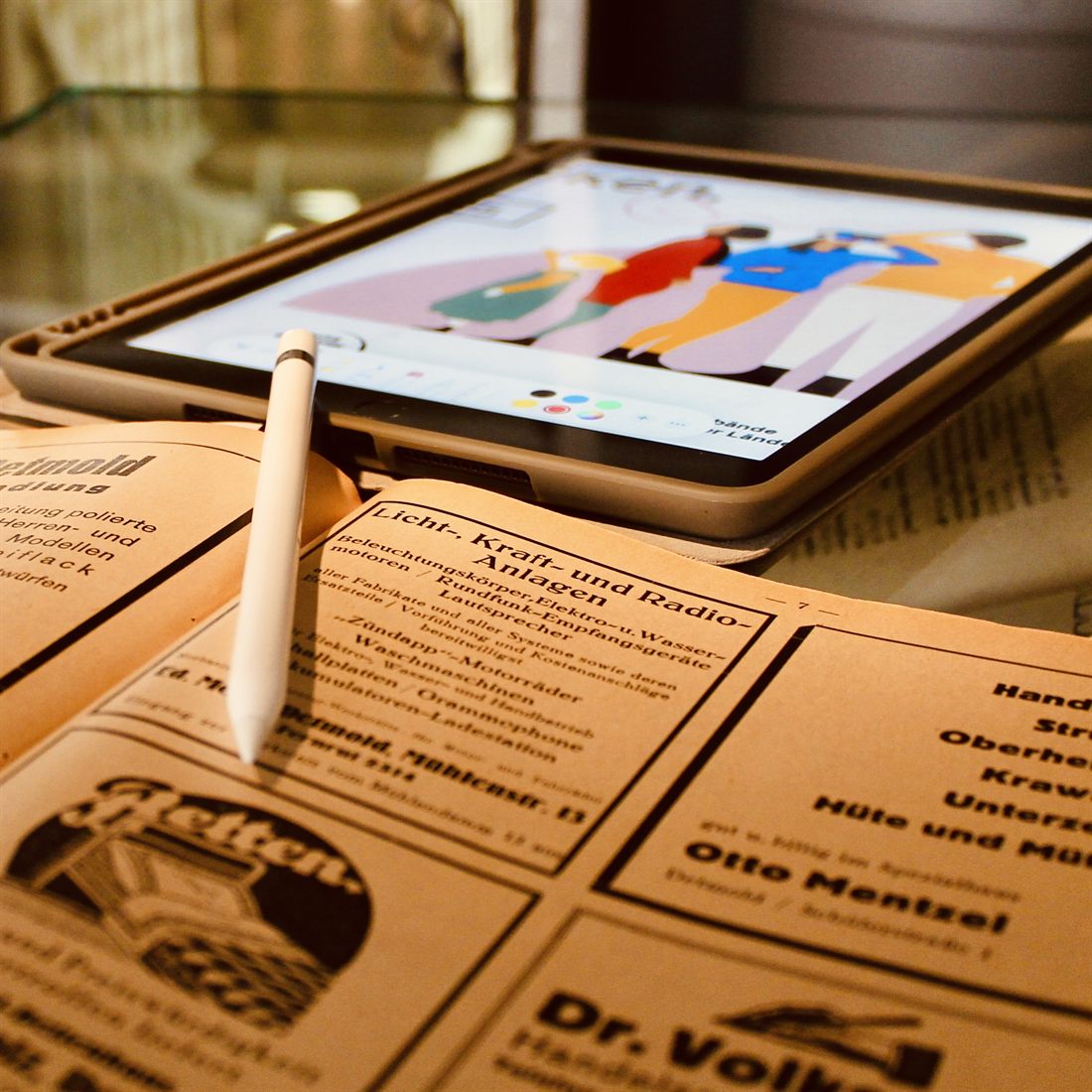 Historische Zeitungsanzeigen auf bräunlichem Papier, dahinter ein Tablet mit einer bunten digitalen Zeichnung von drei Figuren, davor ein Eingabestift.