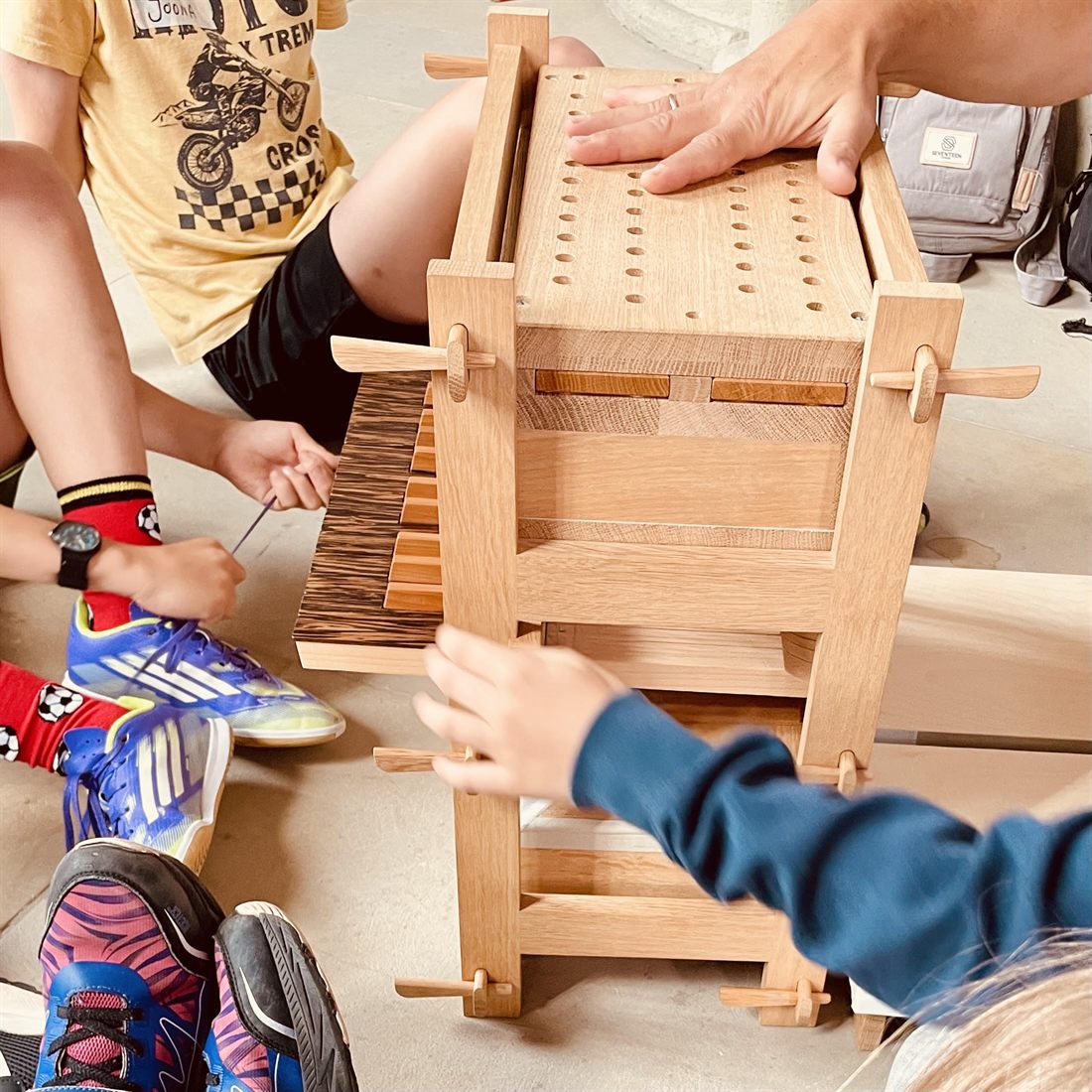 Ein Modell einer Orgel aus hellem Holz steht auf dem Boden, Kinder sitzen darum herum. Eine Hand liegt auf der gelochten Deckplatte, Kinderhände greifen nach der Klaviatur aus Holzstäben.